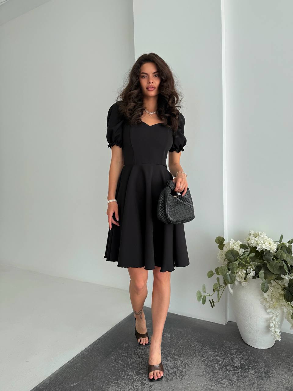 Woman in a black dress holding a black handbag in an indoor setting with white walls and a vase of flowers.