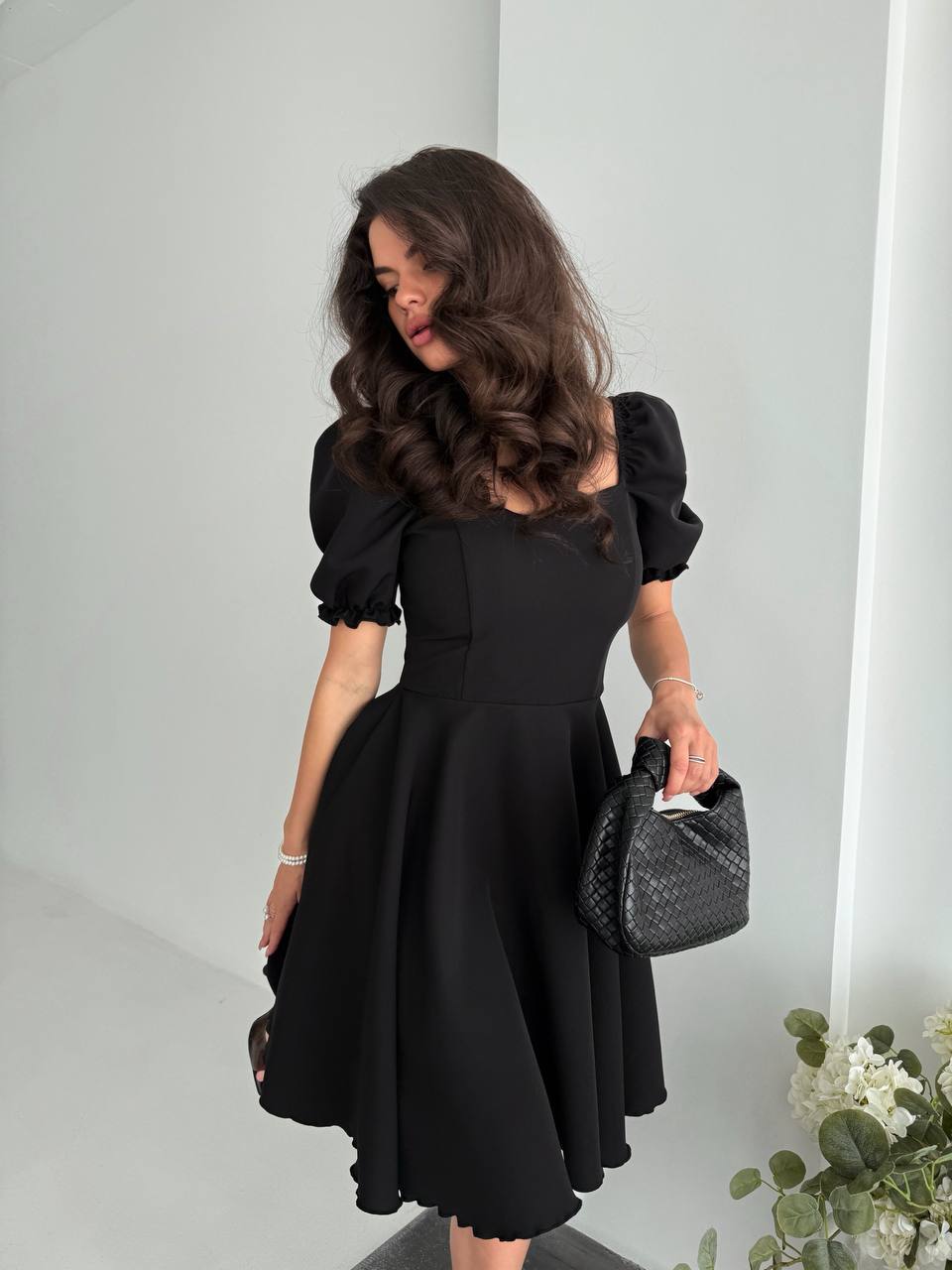 Woman in a black dress holding a black handbag against a white wall.