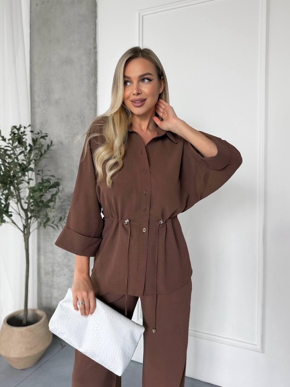 Woman in a brown outfit standing indoors with a white wall and plant in the background