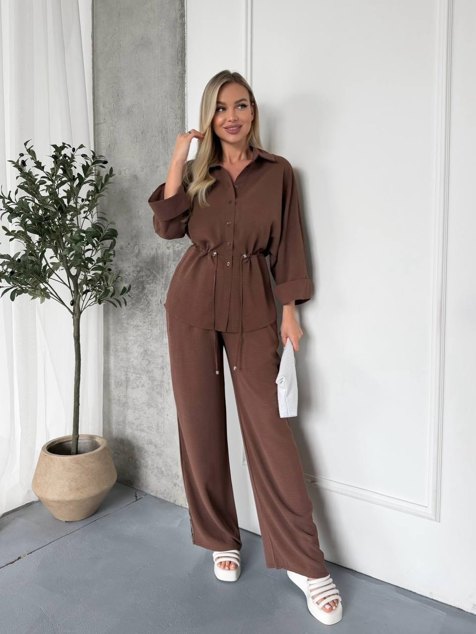 Woman in a brown outfit standing next to a plant indoors