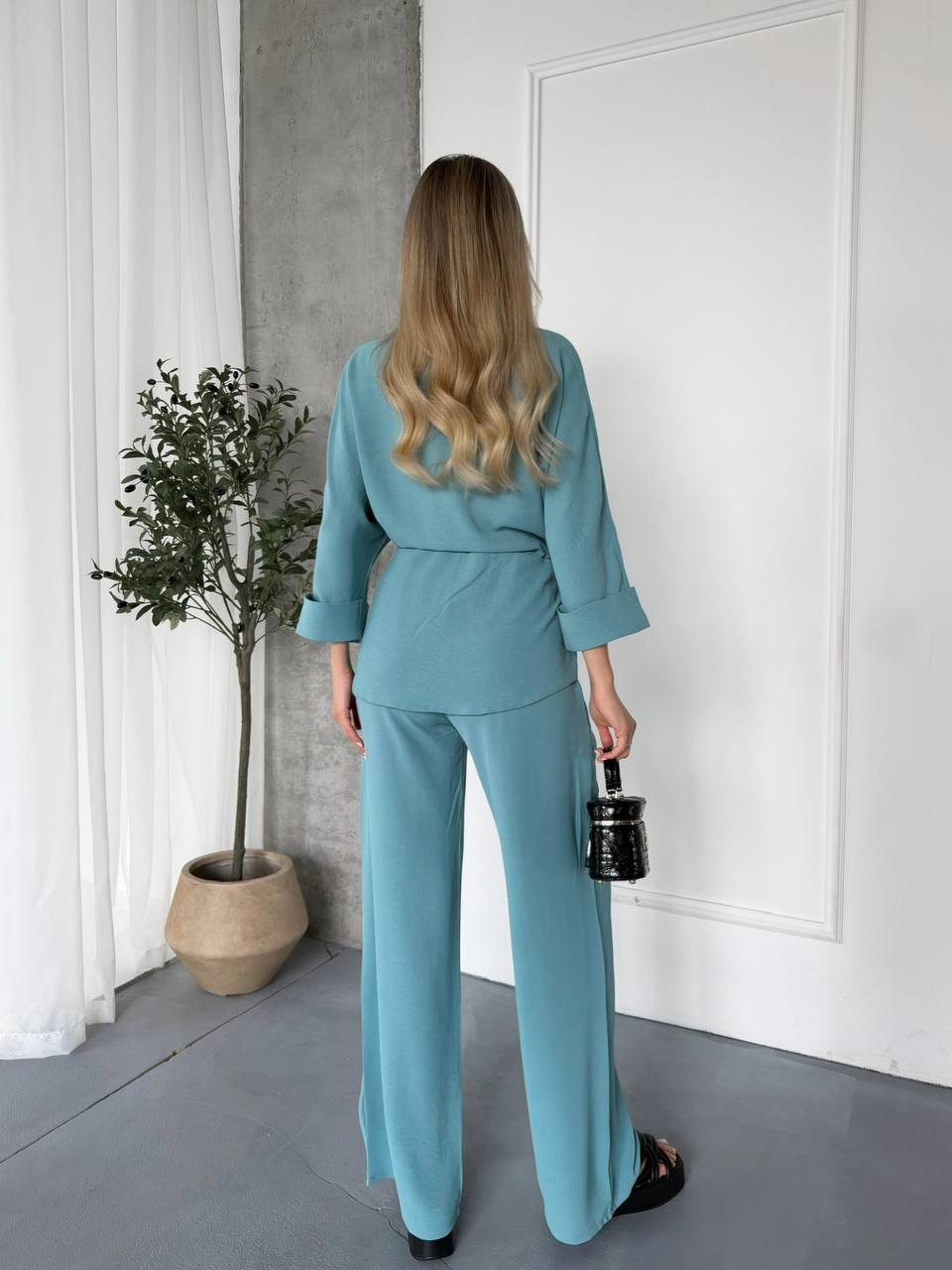Woman in a teal outfit standing in a room with a plant and white walls.