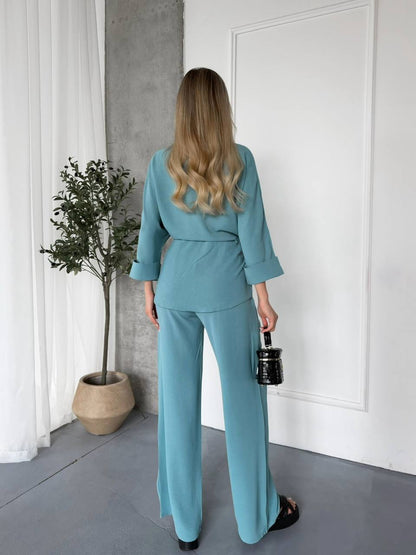 Woman in a teal outfit standing in a room with a plant and white walls.