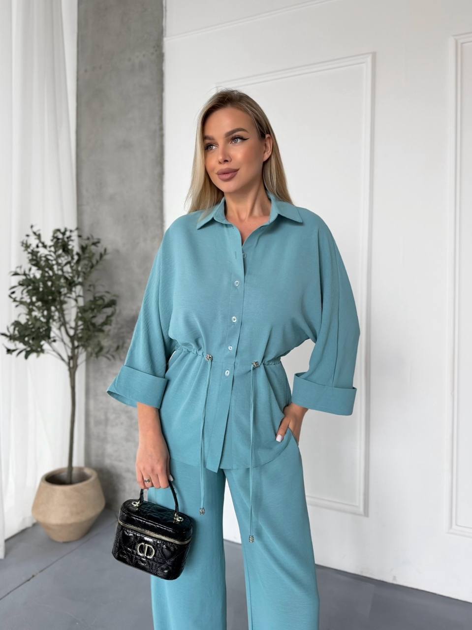 Woman in a teal outfit holding a black handbag indoors.
