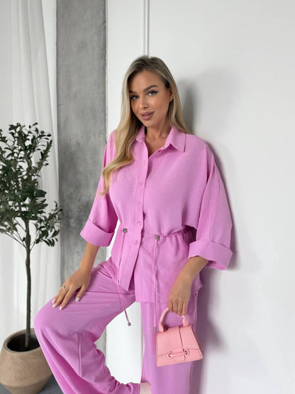 Woman in a pink outfit holding a matching handbag against a white wall.