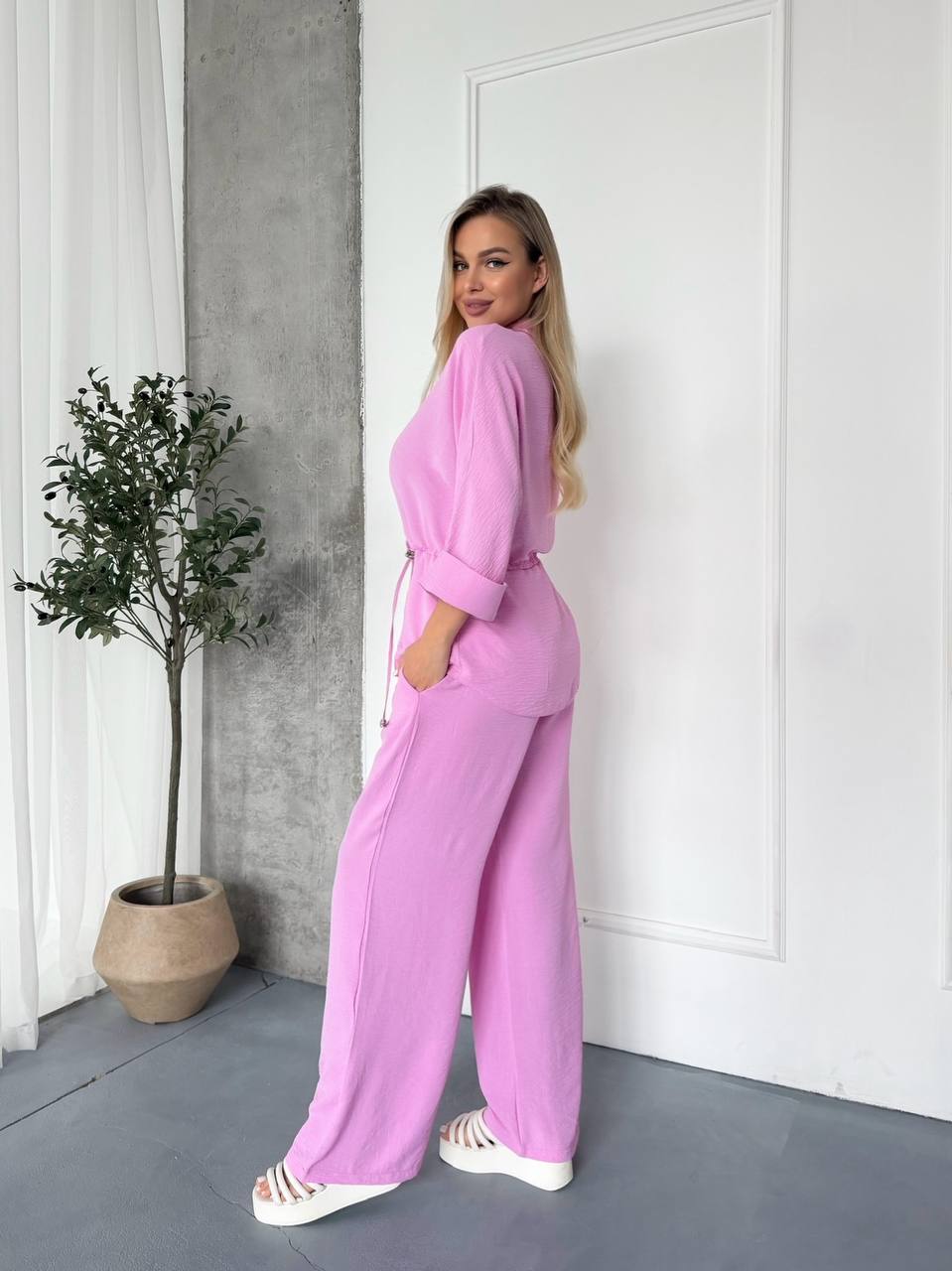 Woman in a pink outfit standing in a room with a plant and white door.
