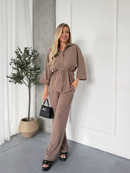 Woman in a brown outfit standing in a room with a plant and white wall.