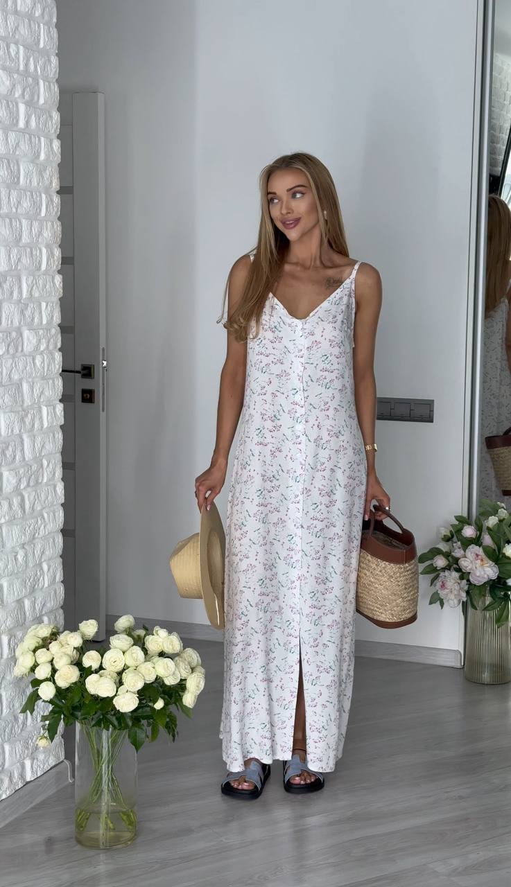 Woman in a white floral dress holding a straw bag and hat, standing in a room with flowers and a textured wall.