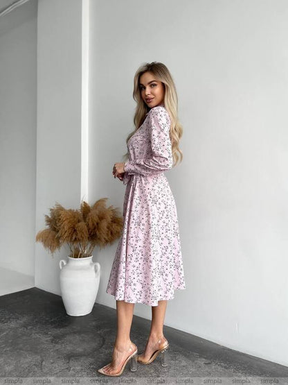 Woman wearing a floral dress standing against a white wall with a vase of dried plants.