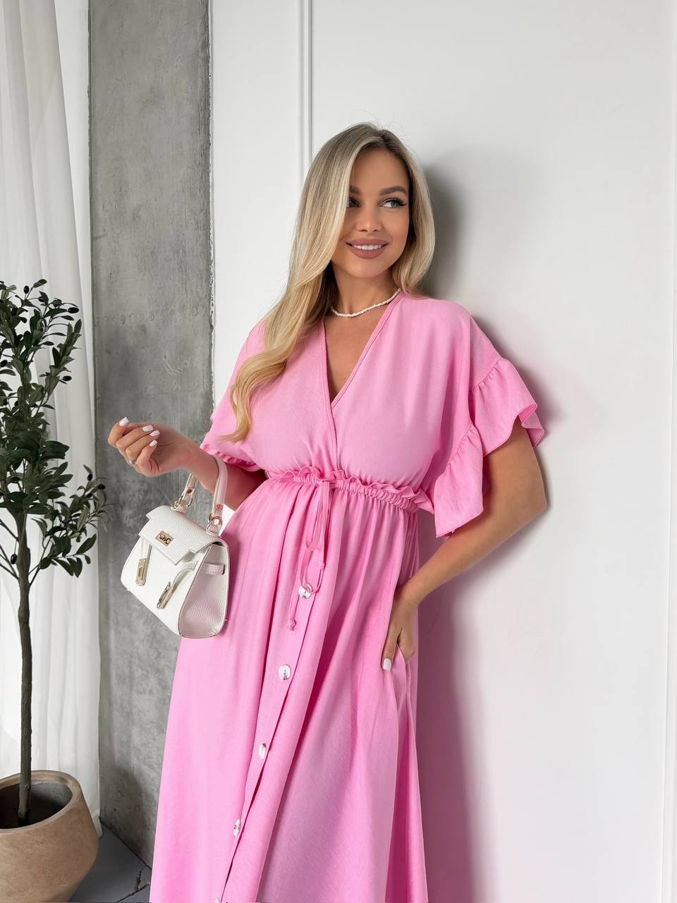 Woman in a pink dress standing against a white wall with a plant to her left.