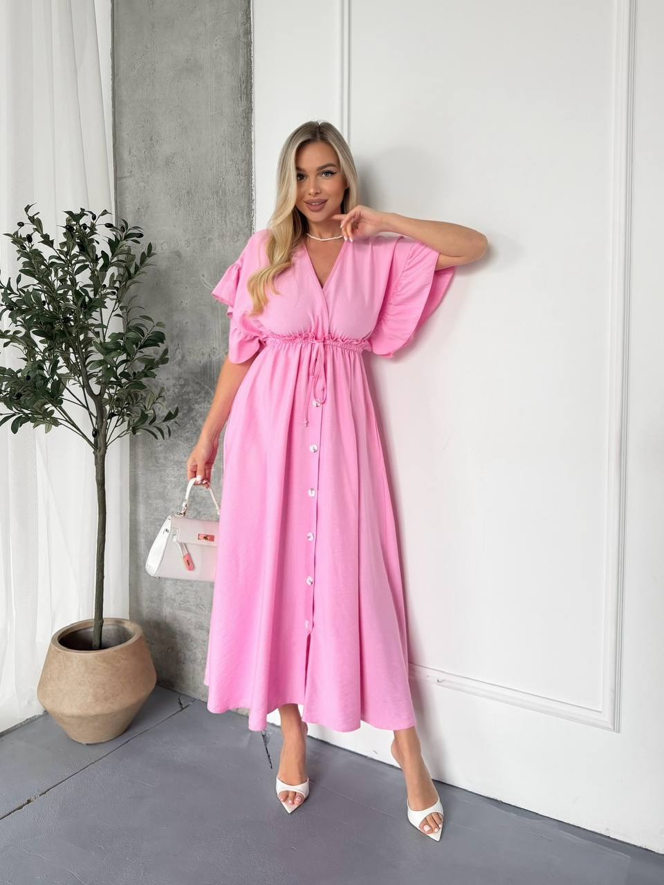 Woman in a pink dress standing against a white wall with a plant to her left.