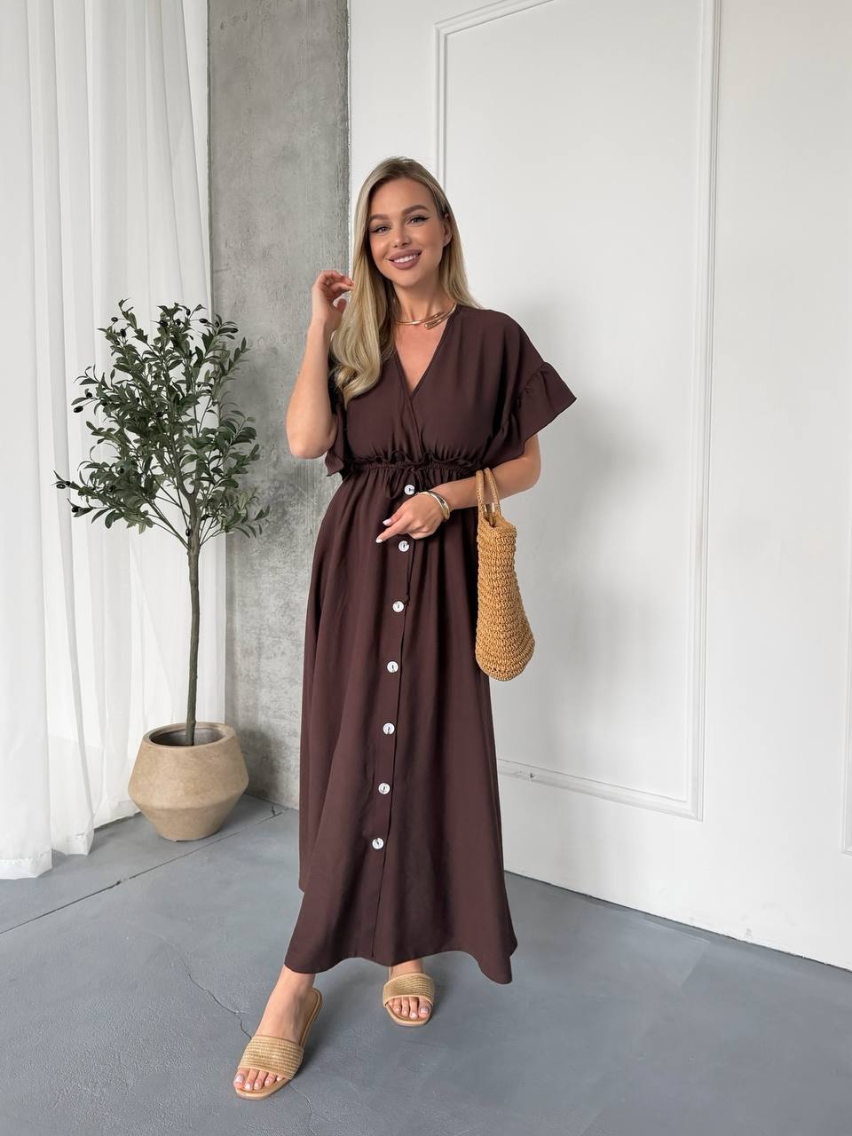 Woman wearing a brown dress standing indoors with a plant and white curtain in the background