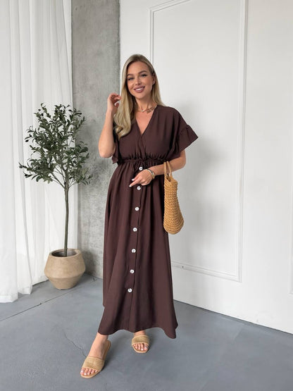 Woman wearing a brown dress standing indoors with a plant and white curtain in the background