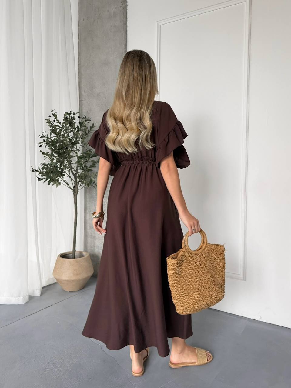 Woman in a brown dress holding a woven bag in a minimalistic room.