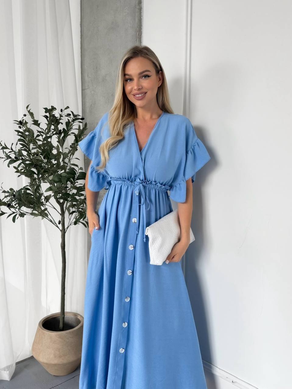 Woman wearing a blue dress standing indoors with a plant and white curtain in the background