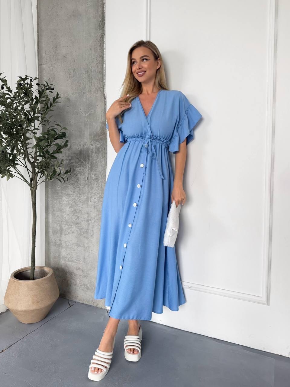 Woman wearing a blue dress standing indoors next to a plant.