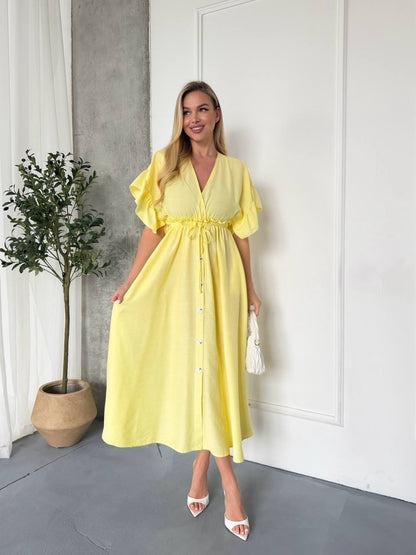 Woman wearing a yellow dress standing indoors with a plant and white wall in the background