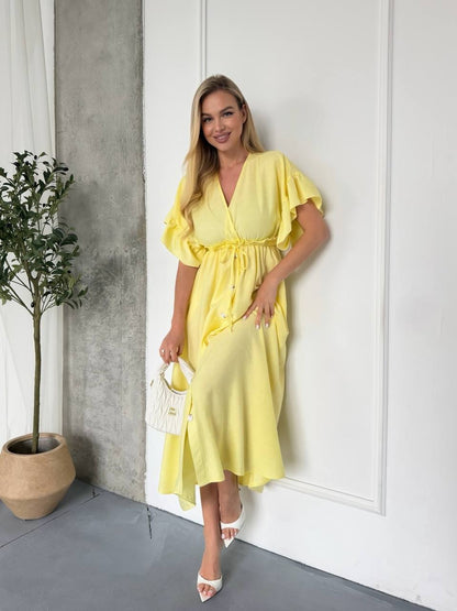 Woman in a yellow dress standing against a white wall with a plant to her left.