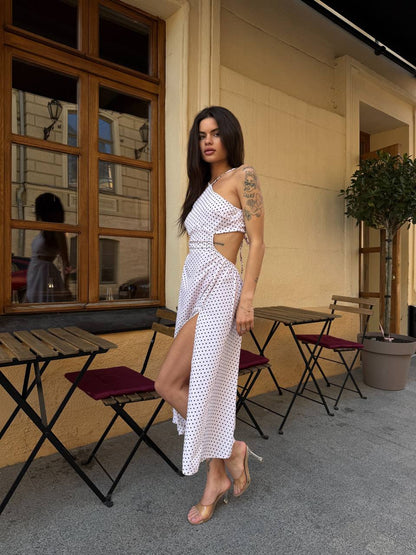 Woman in a white polka dot dress standing on a street with tables and chairs in the background.