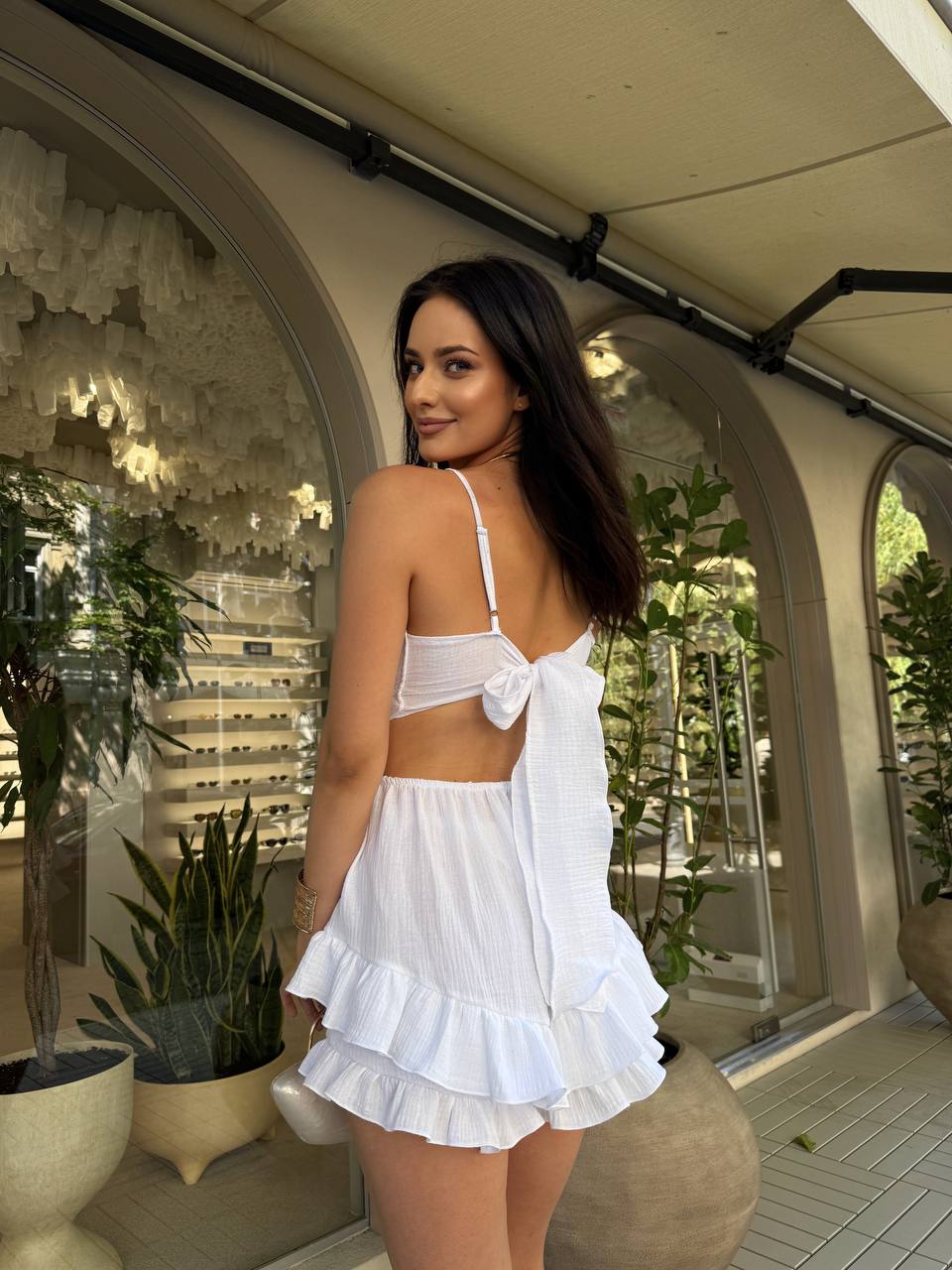 Woman in a white outfit standing in an outdoor setting with plants and architectural elements.