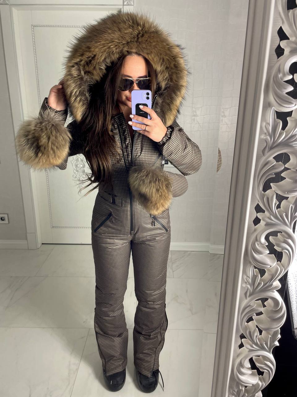 Person wearing a fur-lined hooded jacket and snow pants in front of a mirror.