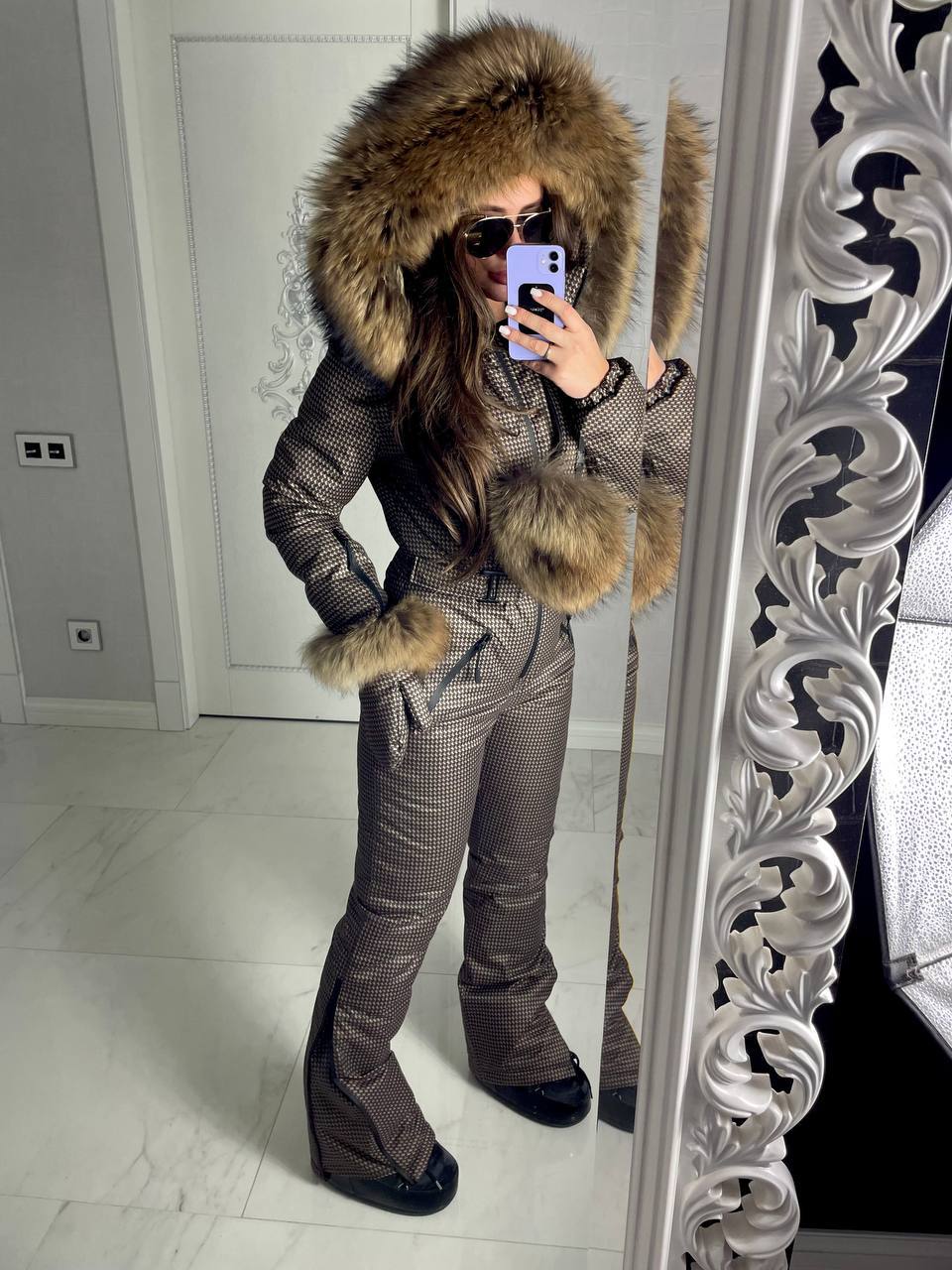 Person wearing a checkered snowsuit with a fur-lined hood in front of a decorative mirror.