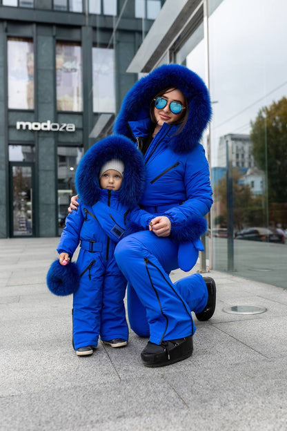 Woman and child in matching blue snowsuits standing on a city street.