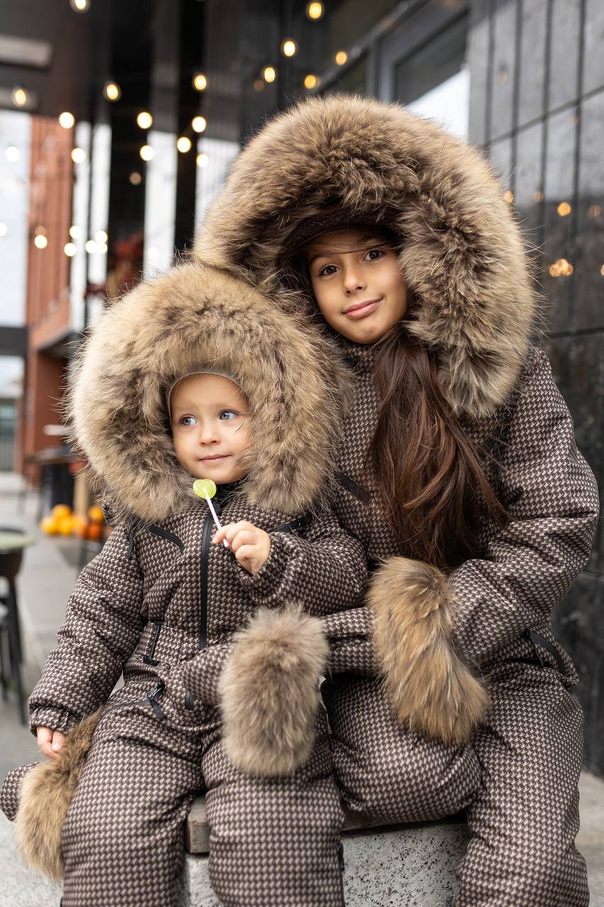 Two children in matching brown winter coats with fur hoods sitting together outdoors.
