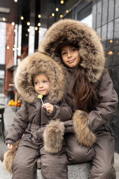Two children in matching brown winter coats with fur hoods sitting together outdoors.