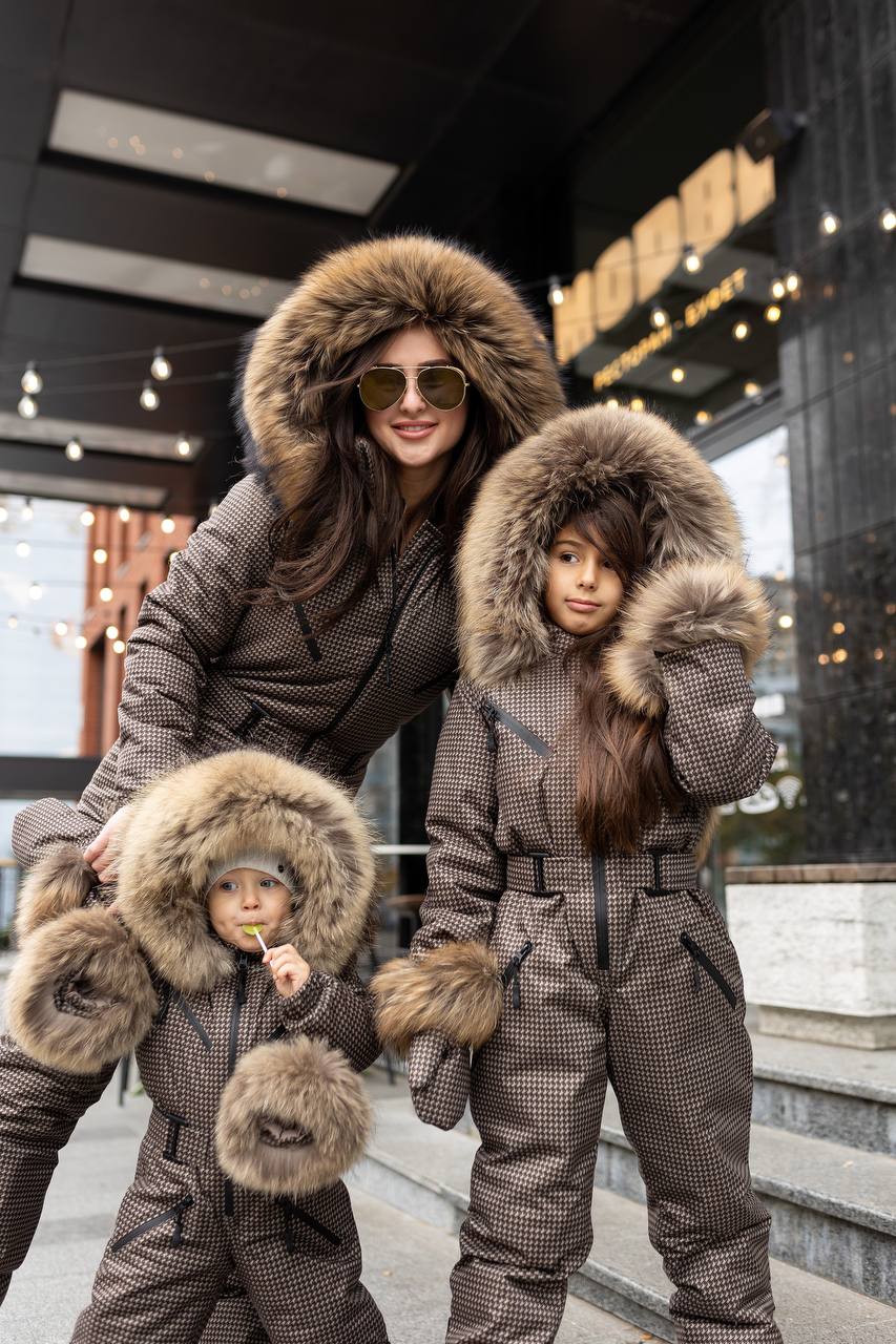 Two children and a woman wearing matching brown winter coats with fur hoods in an urban setting.