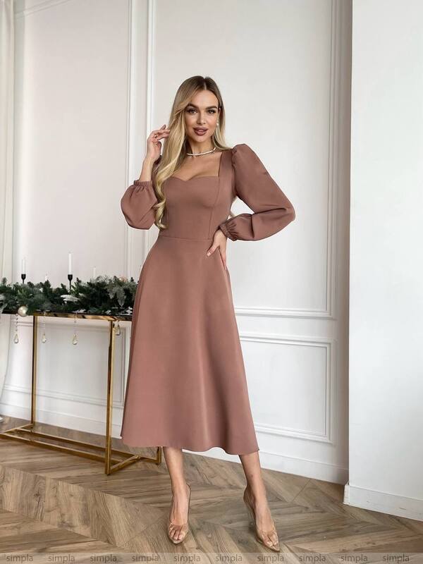 Woman wearing a brown dress standing in a room with a decorative table and wall.