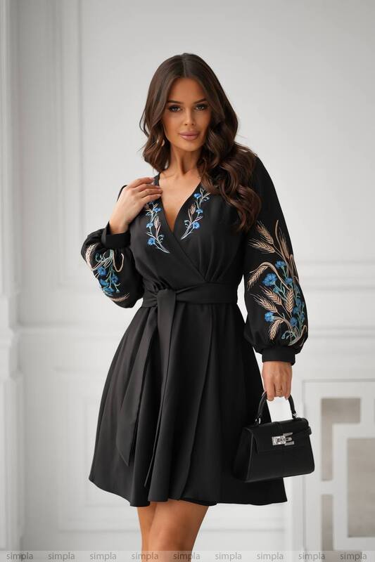 Woman wearing a black dress with floral embroidery holding a black handbag against a white background