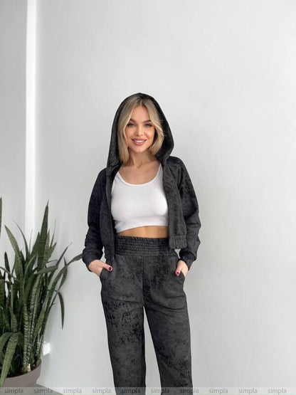 Woman wearing a black hoodie and dark pants standing against a white wall with a plant to the left.