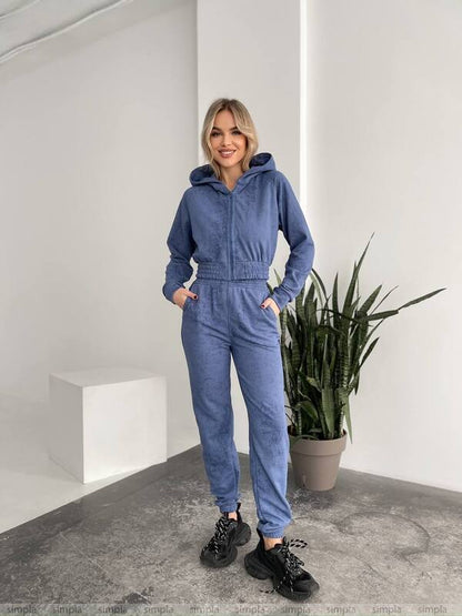 Woman wearing a blue tracksuit standing in a minimalistic room with a plant.