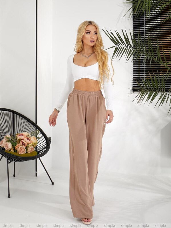 Woman wearing a white top and beige pants in a minimalistic setting with a plant and chair.