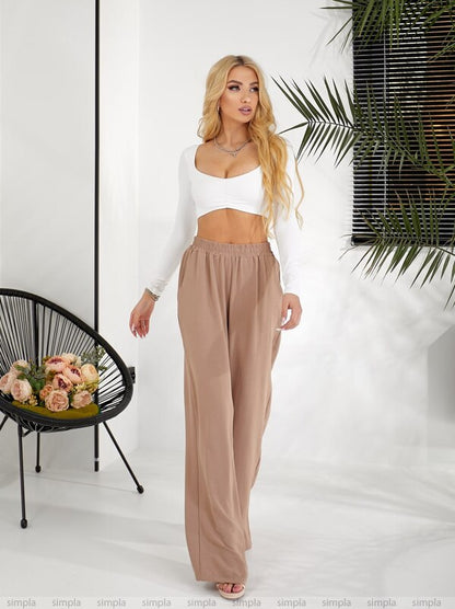 Woman wearing a white top and beige pants in a minimalistic setting with a plant and chair.