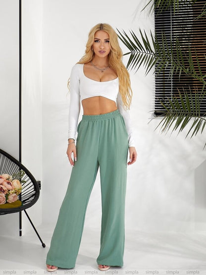 Woman wearing a white top and green pants against a white background with a plant.