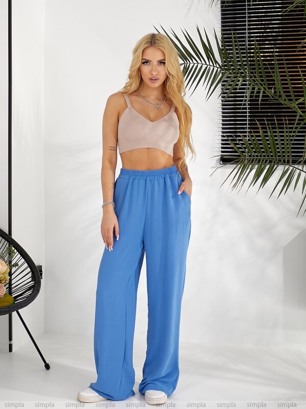 Woman wearing a beige top and blue pants standing against a white wall with a plant in the background.