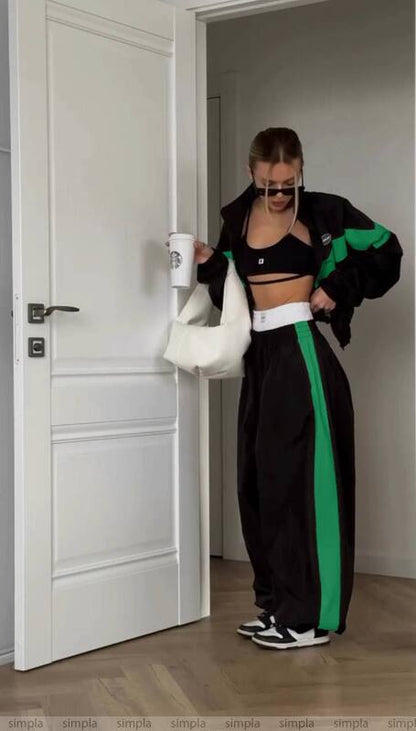 Person in black and green tracksuit with white sports bra and leggings, holding a white bag in a room.