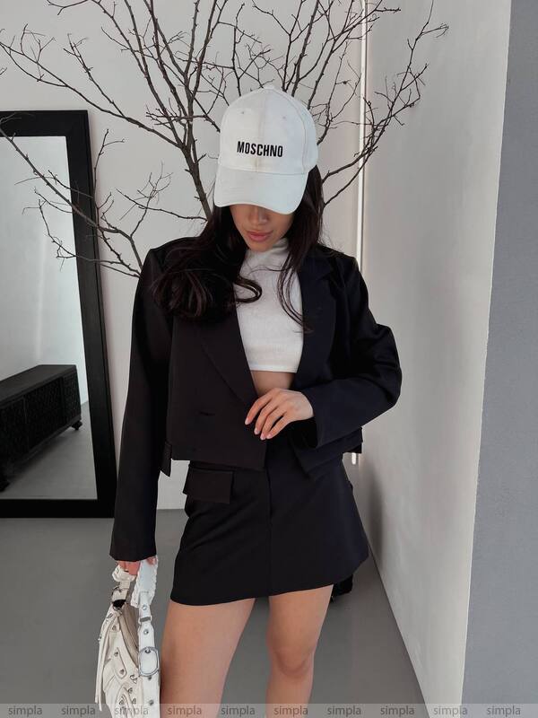 Person wearing a black blazer and skirt set with a white cap indoors.