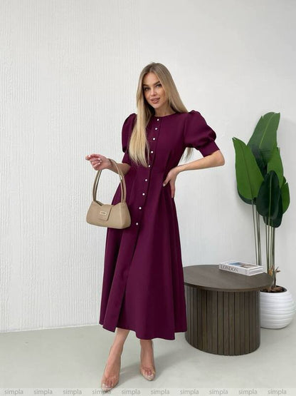 Woman in a burgundy dress holding a beige handbag in a minimalistic room.