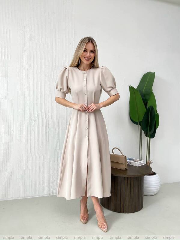 Woman in a beige dress standing in a minimalistic room with a plant and small table.