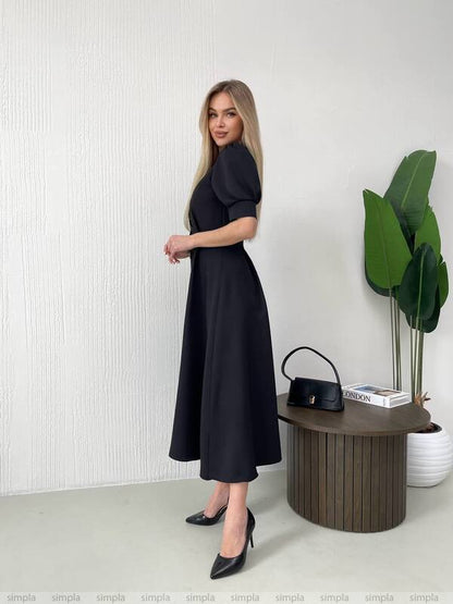 Woman in a black dress standing next to a wooden table with a plant and handbag.