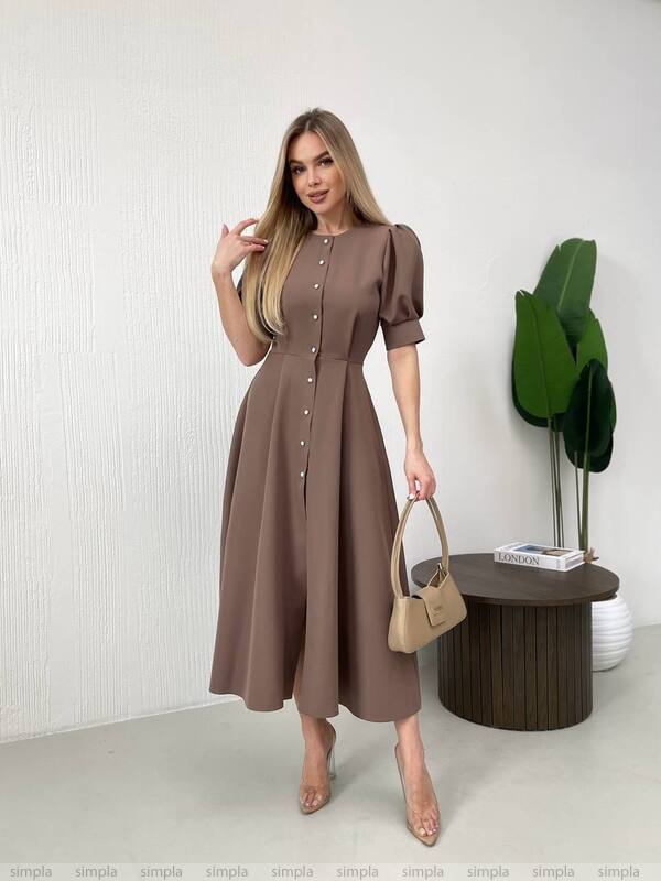 Woman wearing a brown dress holding a beige handbag in a minimalistic setting.