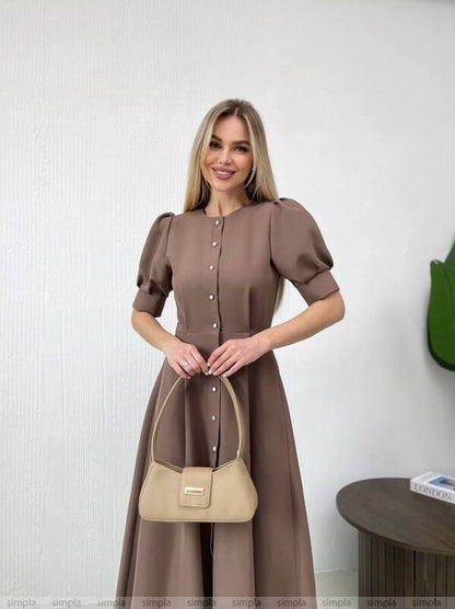 Woman in a brown dress holding a beige handbag against a white wall.