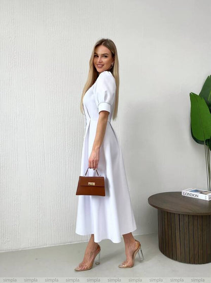 Woman in a white dress holding a brown handbag against a plain background