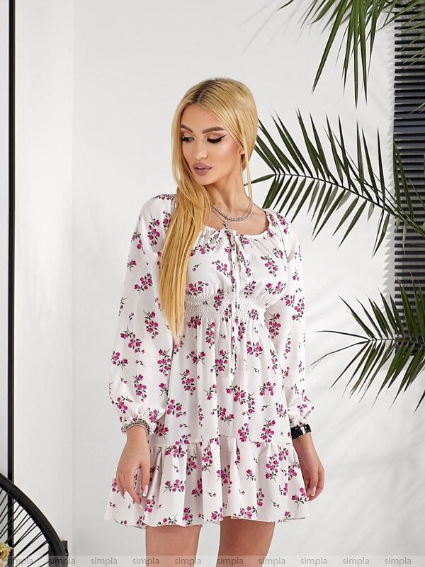 Woman wearing a floral dress standing against a white wall with green leaves.