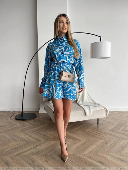 Woman in a blue patterned dress standing in a modern room with a white wall and wooden floor.