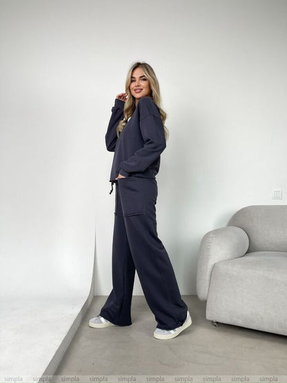 Woman wearing a navy tracksuit standing in a minimalistic room with a gray sofa.