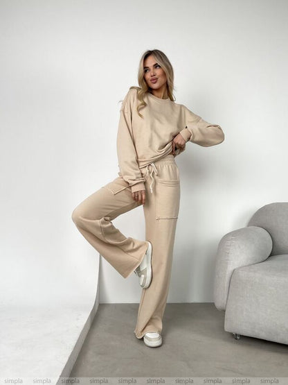 Woman wearing a beige tracksuit standing in a minimalistic room with a gray sofa.