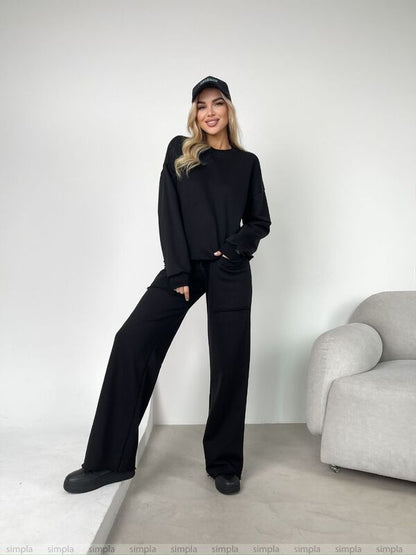 Person wearing a black outfit with a cap in a minimalistic room.